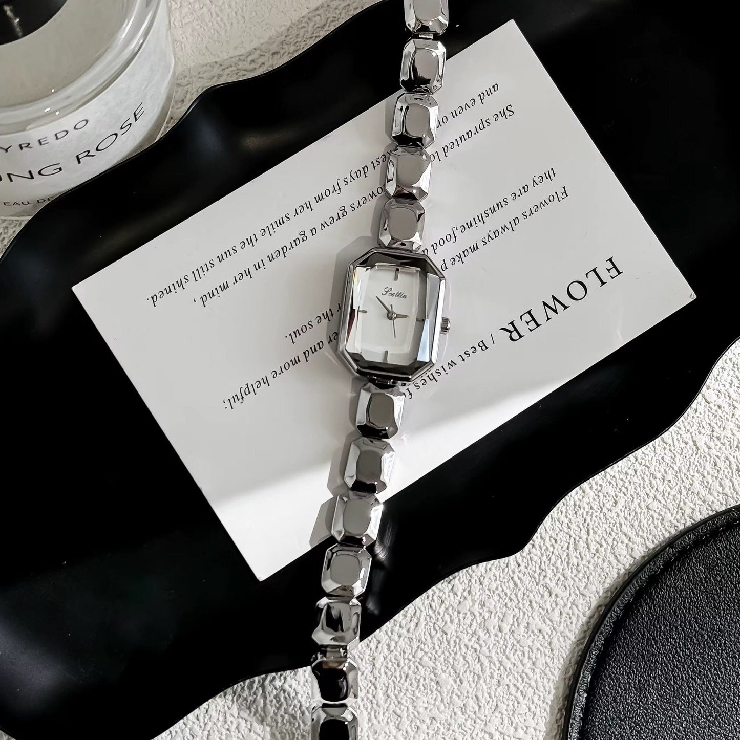 Silver watch with a decorative strap on a white surface with a black tray and container.