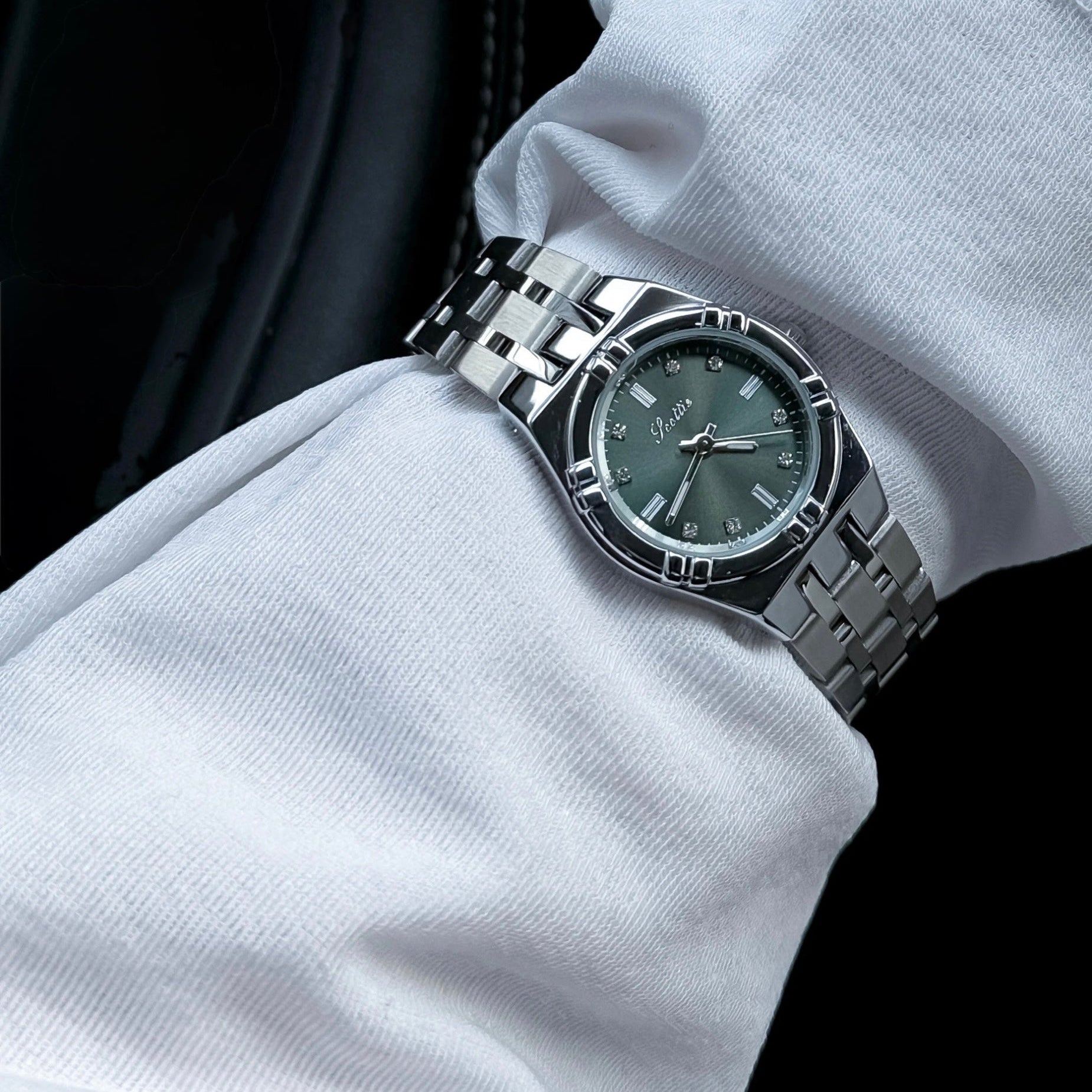 A silver stainless steel wristwatch with a dark green dial and matching green bezel, worn on a person's wrist covered in a white long-sleeve shirt.