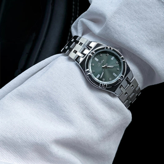 A silver stainless steel wristwatch with a dark green dial and matching green bezel, worn on a person's wrist covered in a white long-sleeve shirt.