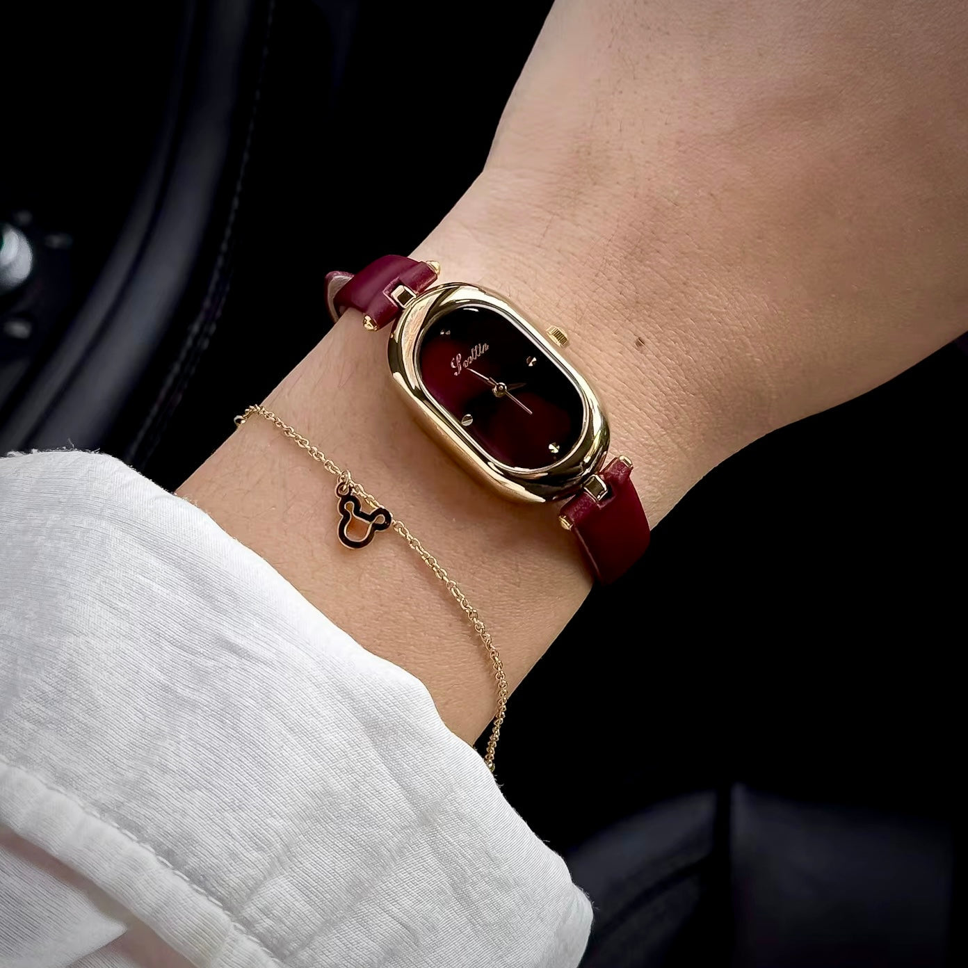 A close-up of a woman's wrist wearing a gold-tone oval watch with a dark red leather strap, alongside a delicate gold chain bracelet, set against a dark background.