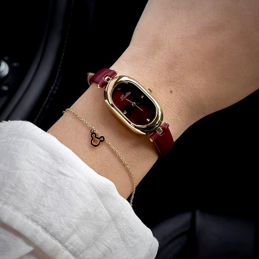 A close-up of a woman's wrist wearing a gold-tone oval watch with a dark red leather strap, alongside a delicate gold chain bracelet, set against a dark background.